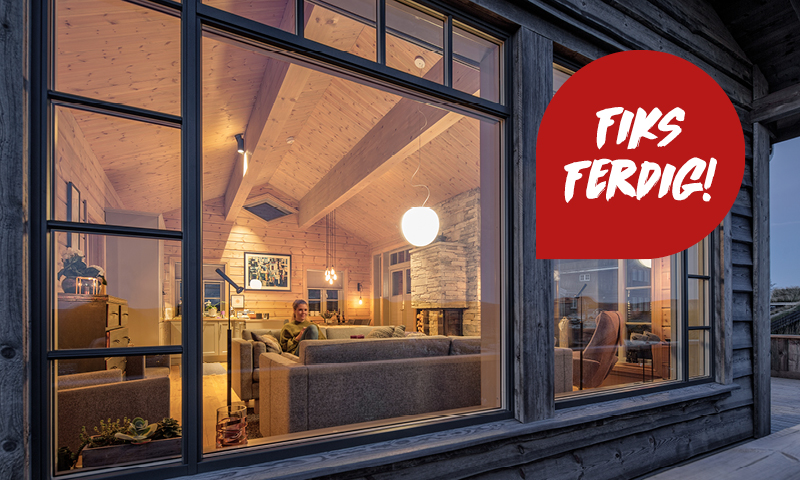 Utvendig vindu mot kveld i trehytte stue med steinpeis, grå sofaer og rødt «Fiks ferdig!» skilt