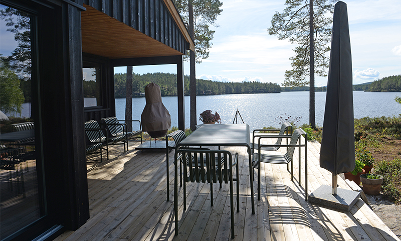 Treterrasse med svart hytte, grønt bord og stoler, sammenbrettet parasoll ved innsjø i solskinn.