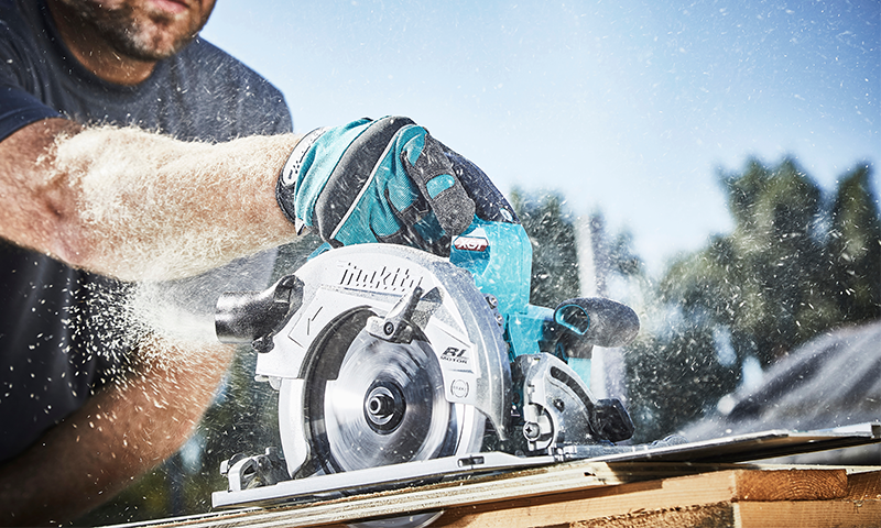 Mann skurer tre utendørs med blå Makita-sirkelsag, sagflis spruter i sollys.