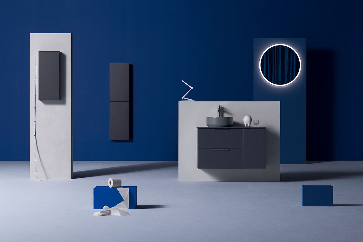 Minimalistisk baderom med blå vegg, grå servantskap, rundt belyst speil og toalettpapir.