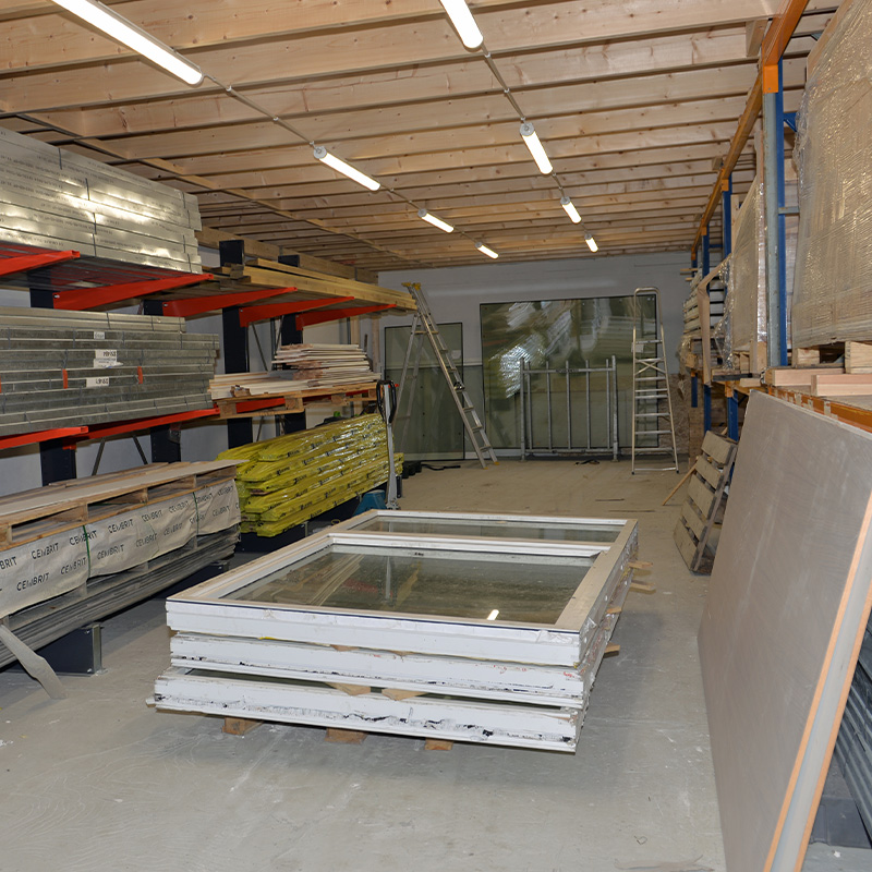 Lagerrom med trehimling, lysrør, stablede hvite vindusrammer, gule isolasjonssekker og metallplater.