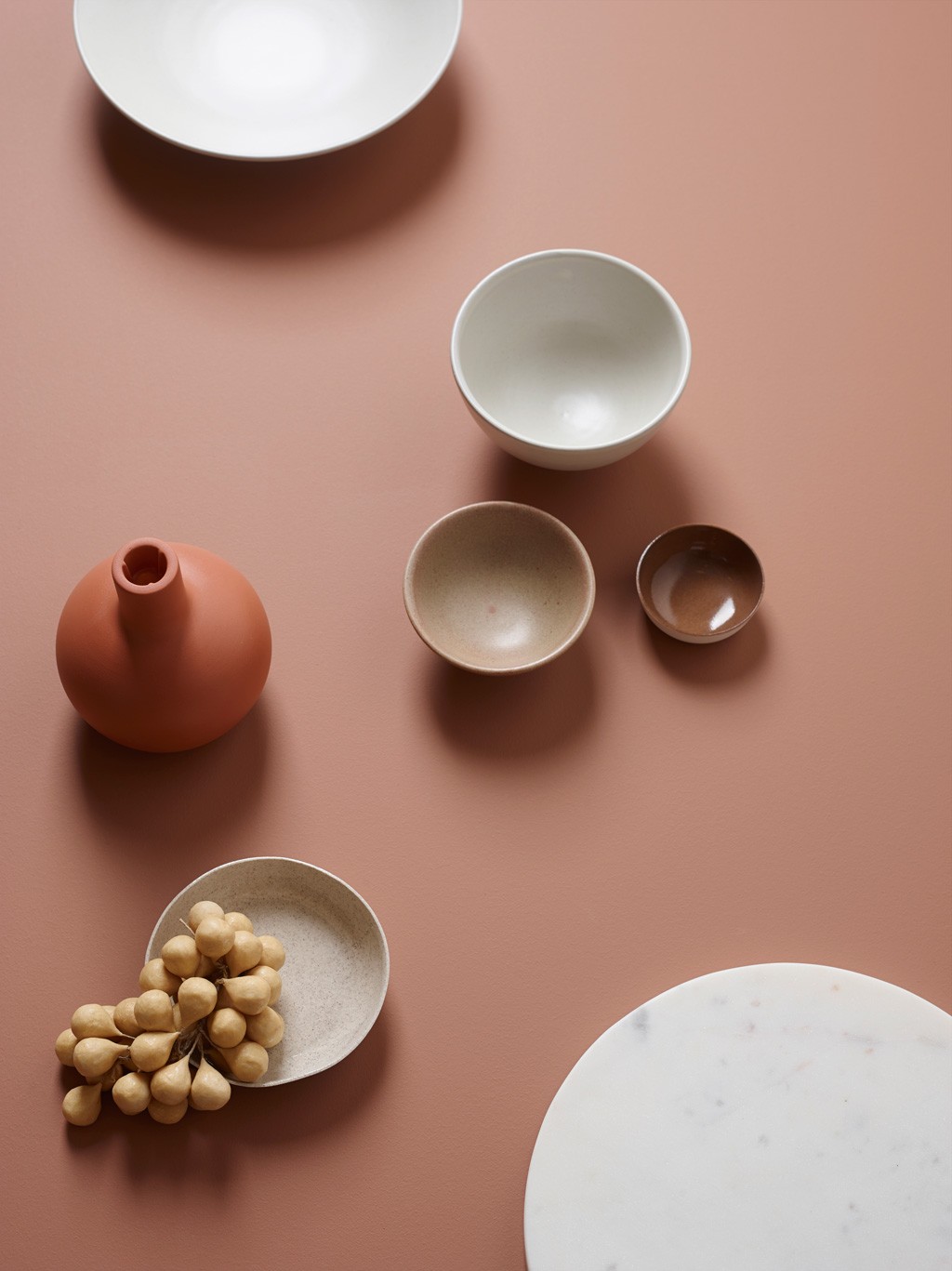 Flatt oppsett med hvite, beige og brune boller, terrakottavase og gule bær på rustfarget bakgrunn.