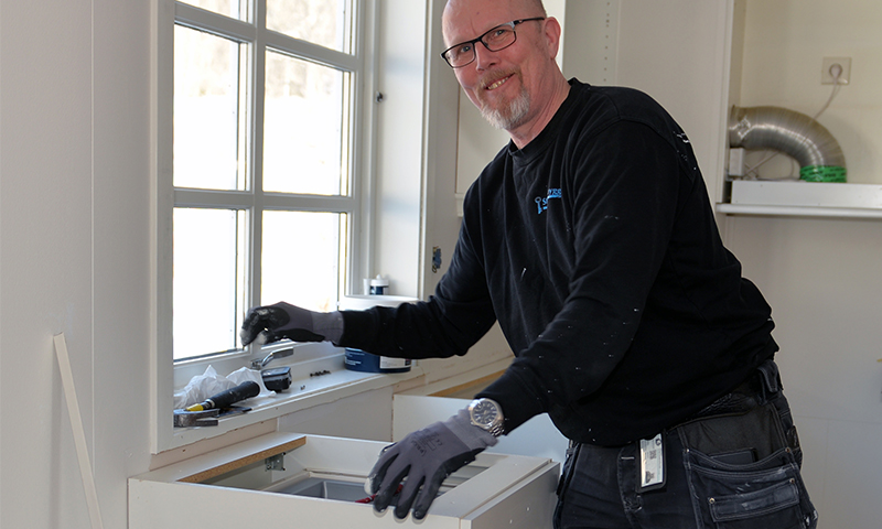 Håndverker i svart genser og grå hansker monterer hvitt kjøkkenskap under vindu.