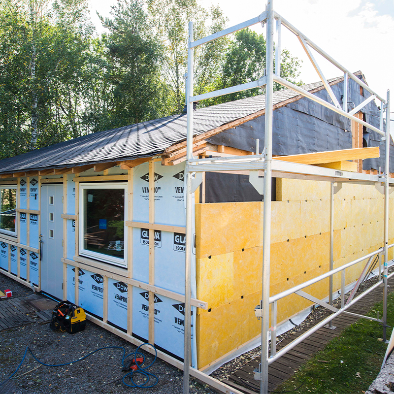 Hus under oppussing med blå duk, gule plater, sølv stillas og grønn skog i bakgrunnen