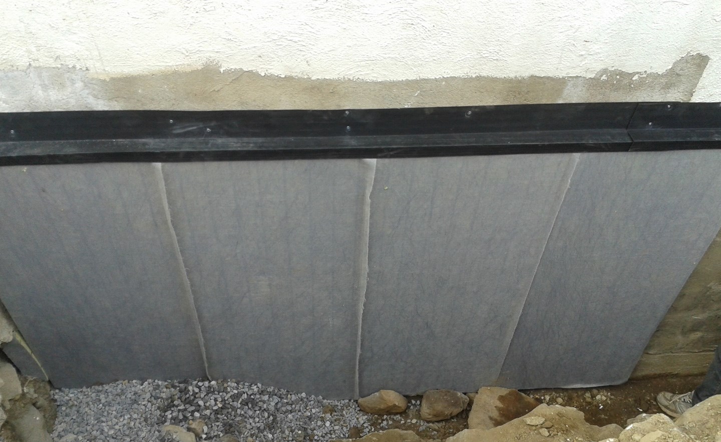 Lysegrå dreneringsplater mot hvit grunnmur, svart topplist og grå pukk nederst.