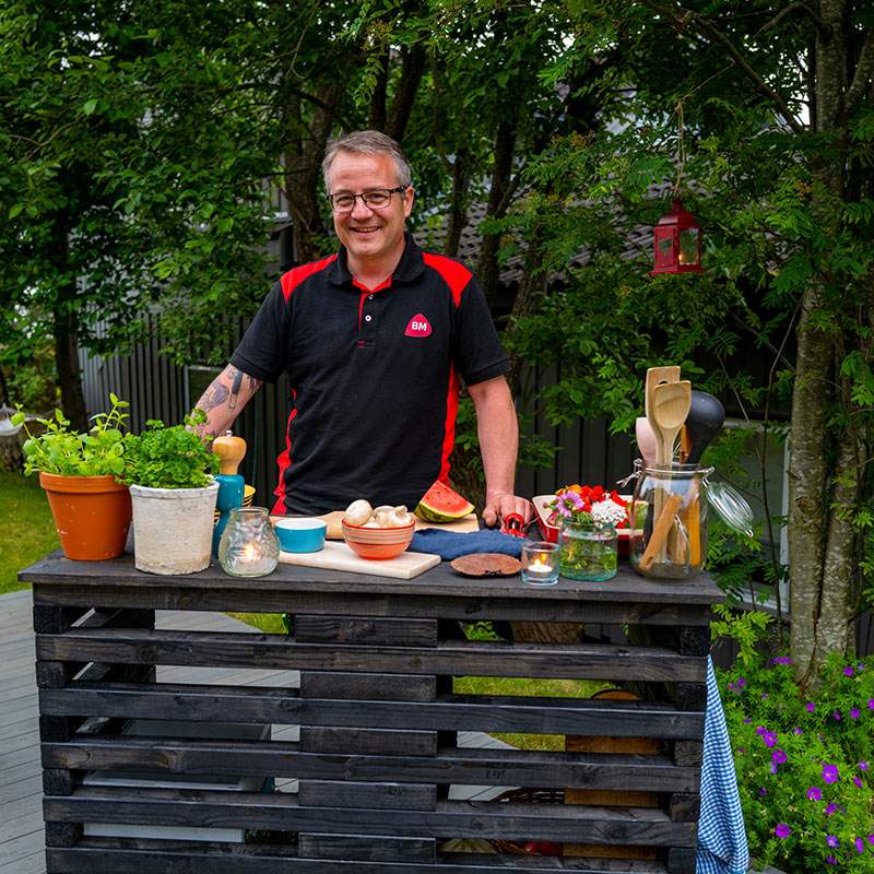 Person ved svart pallebord i hage med grønne planter, melon, sopp og kjøkkenredskaper