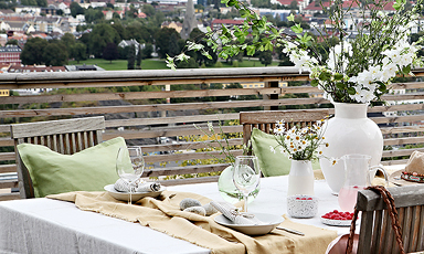 Utebord på terrasse med hvit duk, grønne puter, vaser med hvite blomster og byutsikt
