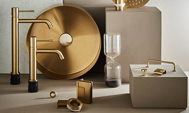 Gylne messingkraner, rund servant, timeglass og avløpsdeksler på beige underlag.