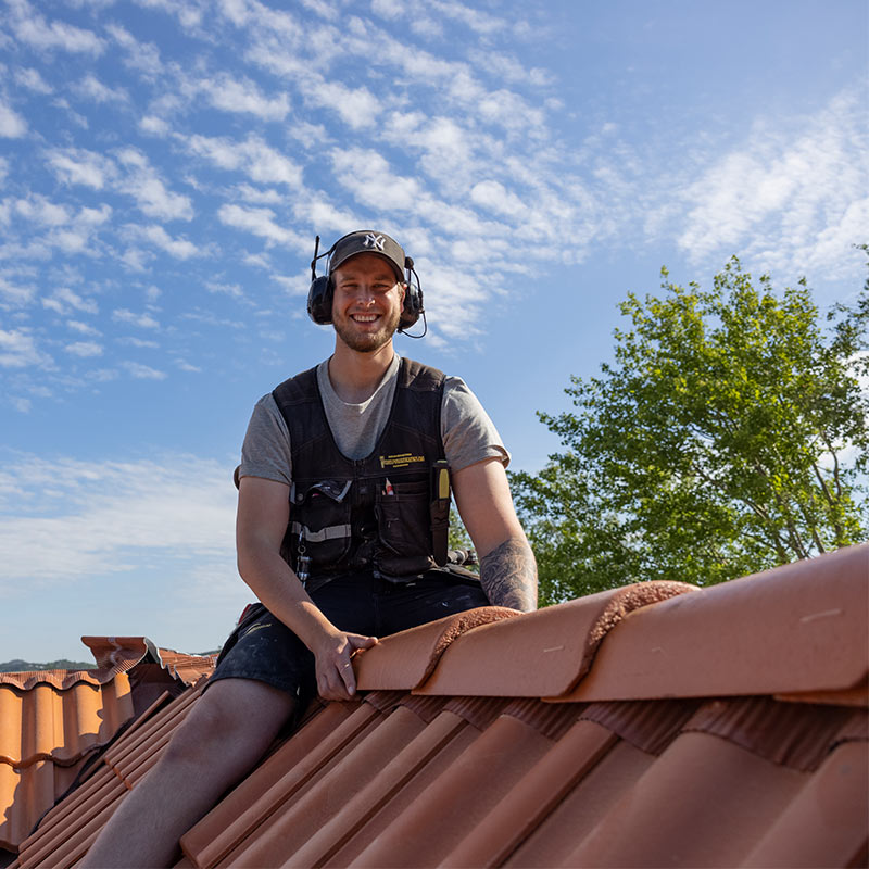 Taktekker i vest på rød takstein under blå himmel fester mønepanne ved grønt tre