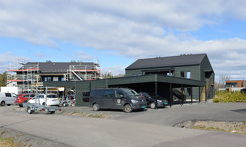 To mørkegrønne boligbygg under oppføring med stillas, biloppstilling og skyet himmel.