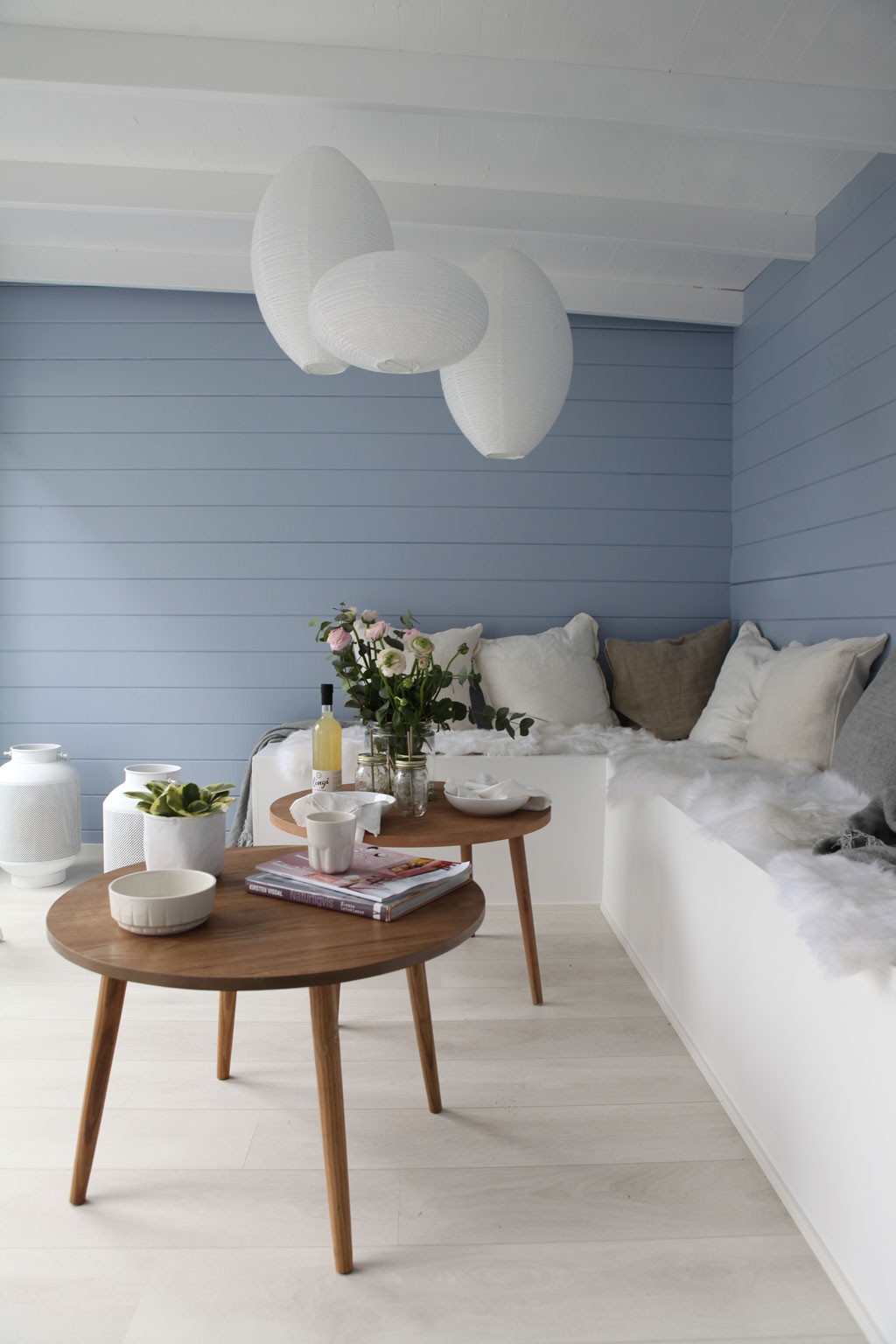 Lyst hjørne med blå panelvegg, hvite papirlykter i taket og to små runde trebord med blomster.