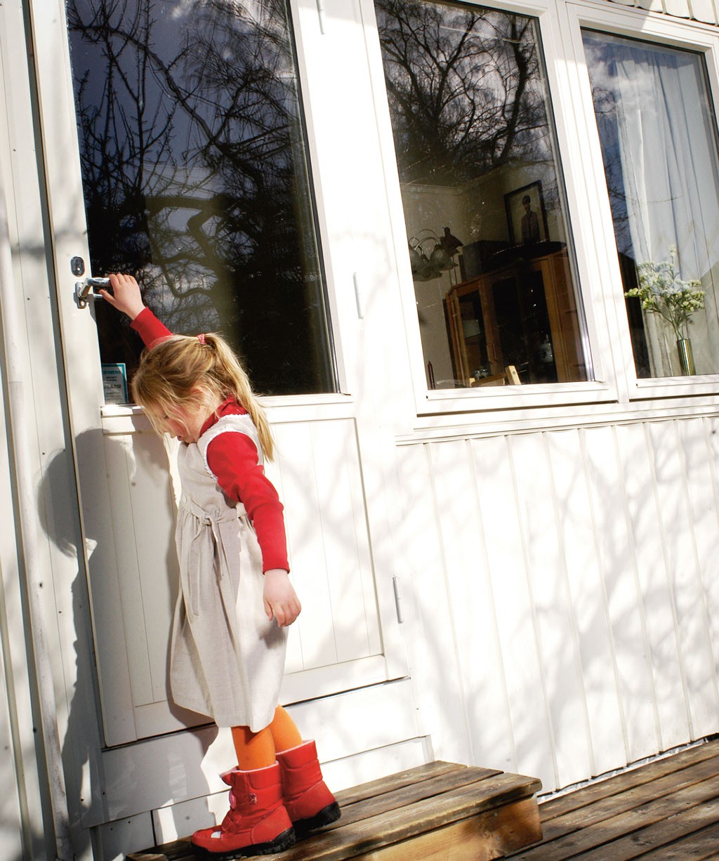 Lite barn i røde støvler åpner hvit dør på treveranda, trær reflektert i vindu.