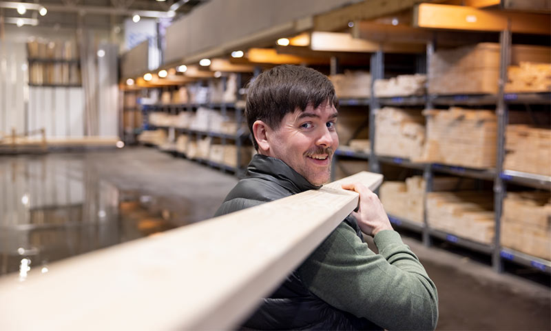 Mann med grønn genser og svart vest bærer en lang treplanke i et lyst trelastlager