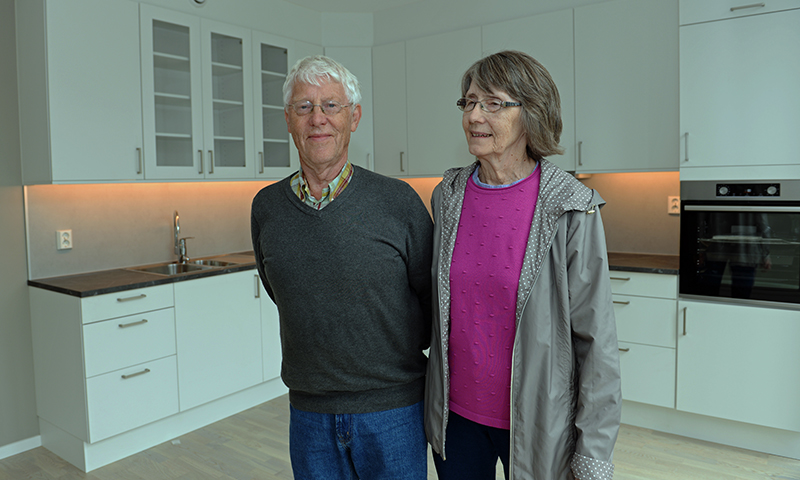 Et eldre ektepar i et moderne hvitt kjøkken med LED-lys, mannen i grå genser, kvinnen i rosa genser.