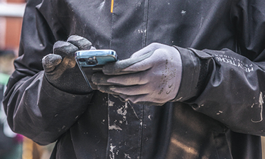 Nærbilde av maler i svart jakke med hvite malingsflekker og grå hansker som bruker mobil.