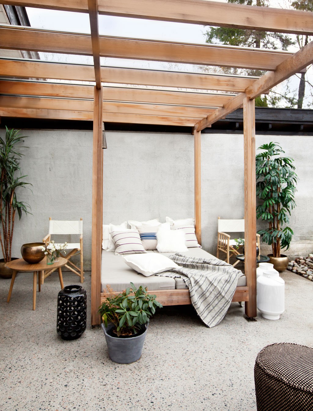 Solfylt uteområde med trepergola, grå dagbed, striperputer, pledd og grønne planter.