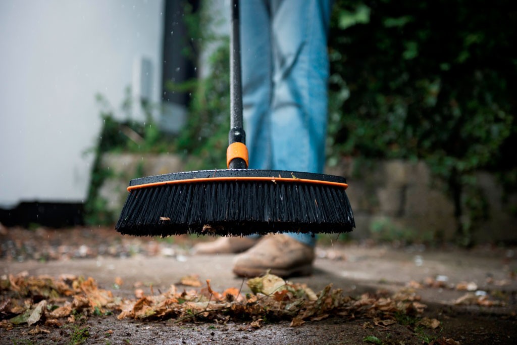 Person i jeans og brune støvler feier våte høstblader med svart kost med oransje kant