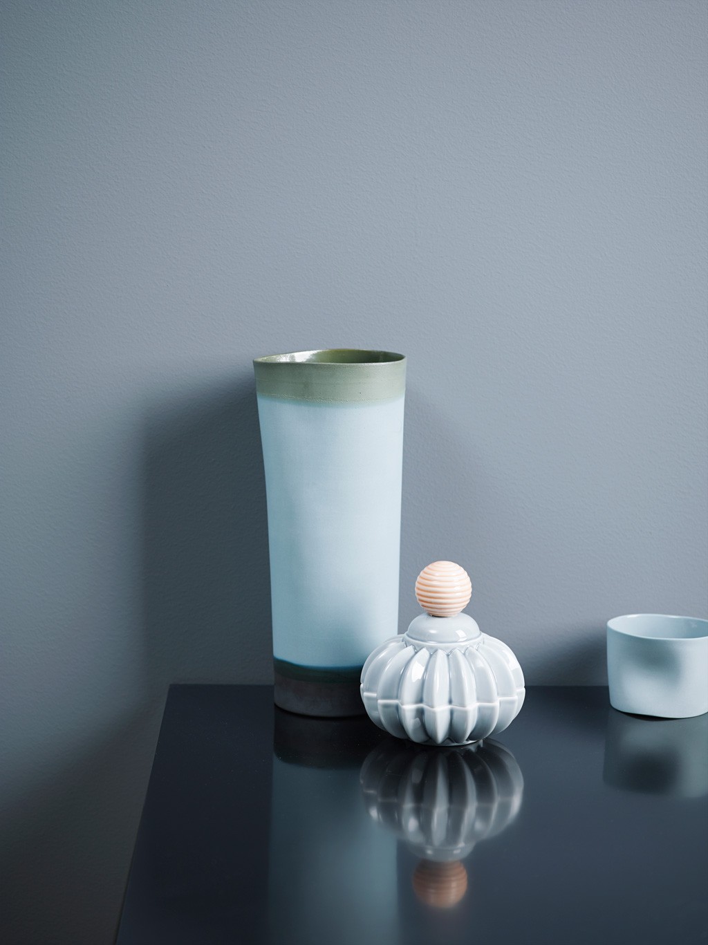 Høy matt keramikkvase (blågrønn), hvit flaske med rosa kulelokk og blå kopp på bord mot grå bakgrunn