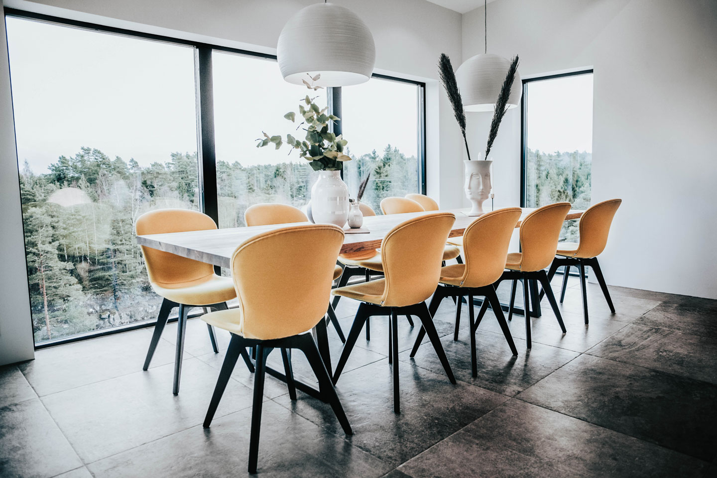 Moderne spisestue med langt trebord, åtte gule stoler, hvite pendellamper og vinduer mot skog.