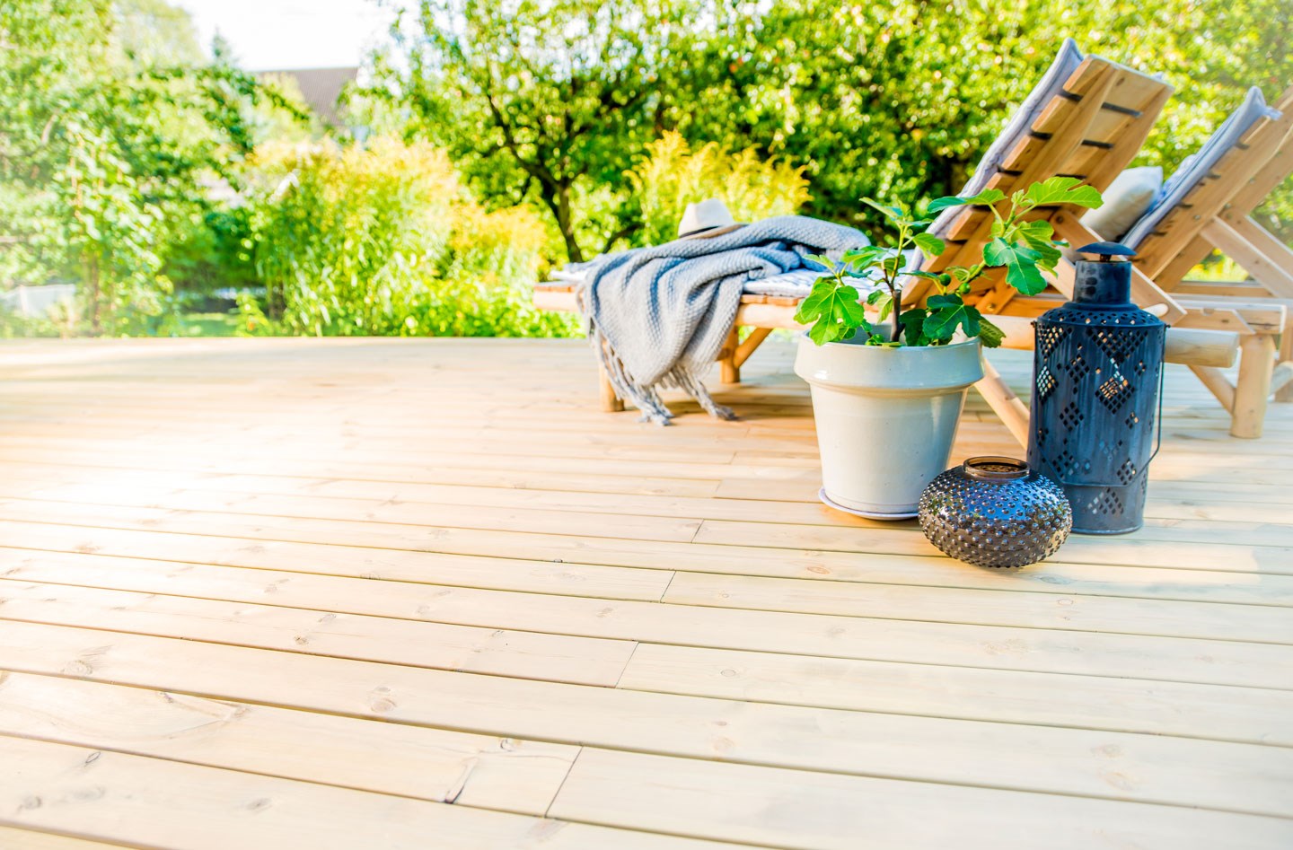 Solfylt treterrasse med to liggestoler, grått pledd, hvit plantepotte, sorte lanterner og grønn hage