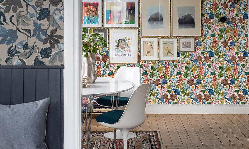 Hvitt rundt bord med hvite stoler og blå seter, fargerik botanisk tapet og gallerivegg.