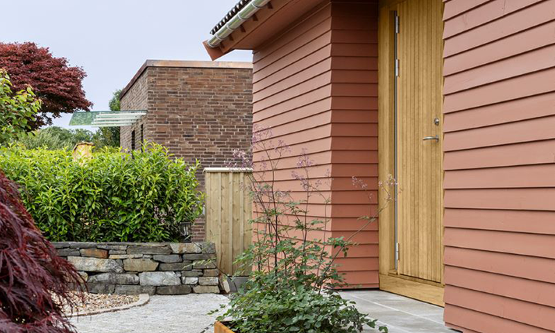 Inngangsparti med rødt trepanel og lys eikedør, foran steinmur, grønn hekk og rød lønn