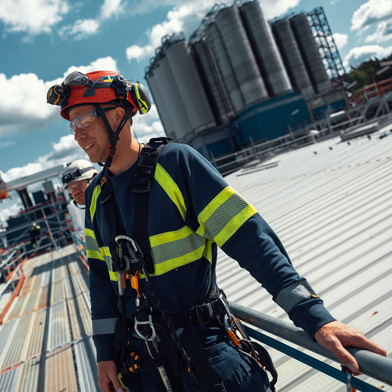 Arbeider i rød hjelm og mørkt tøy med gule reflekser og fallsikring på metalltak ved silotanker