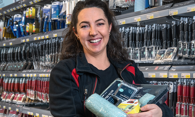 Butikkansatt i svart og rødt holder malingsruller og utstyr foran hylle med malerverktøy