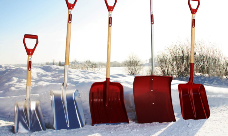 Fem snøspader med røde håndtak, to i aluminium og tre røde, plassert i snø mot vinterhimmel.