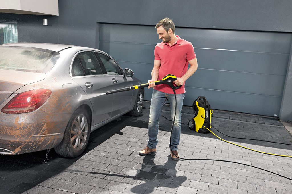 Mann i rød piké vasker gjørmete sølvbil med gul-svart høytrykksvasker på grå innkjørsel.
