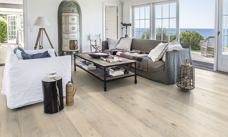 Lyst stue med hvit og grå sofa, rustikt trebord, lyst parkettgulv og havutsikt gjennom store vinduer.