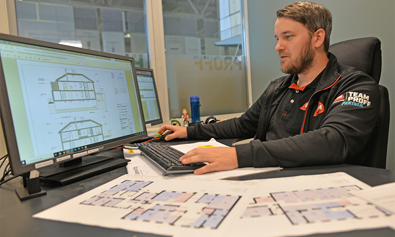 Ansatt i svart Team Proff-jakke ved kontorpult med dataskjerm og papirtegninger av husplaner.