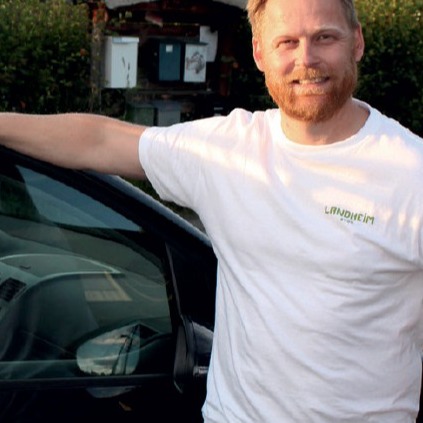 Mann i hvit t-skjorte med grønt logo lener armen på svart bil foran grønn hekk.