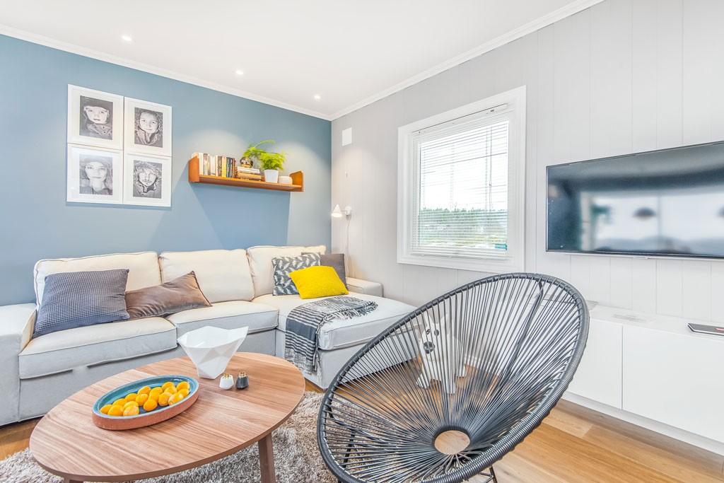 Lys stue med grå sofa og gule puter, blå aksentvegg, trebord med mandariner og svart stol.