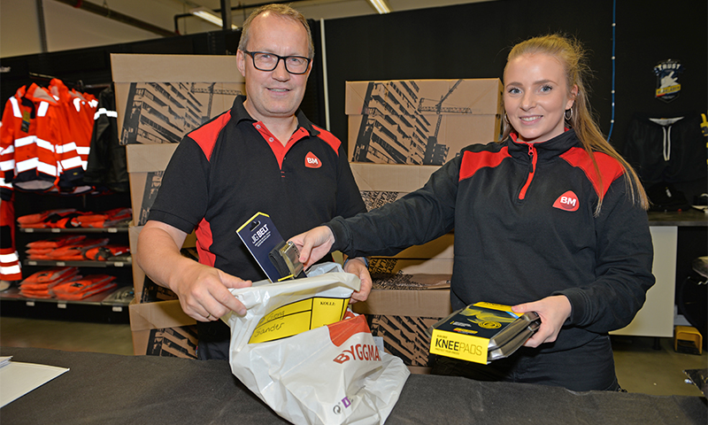 To ansatte i svart-røde klær pakker gule knebeskyttere i hvit plastpose bak disken.