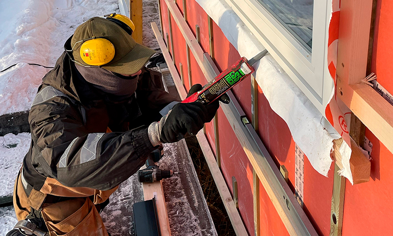Bygningsarbeider i vinterklær med ørevern fuger hvit vinduskarm på rødt hus i snø.