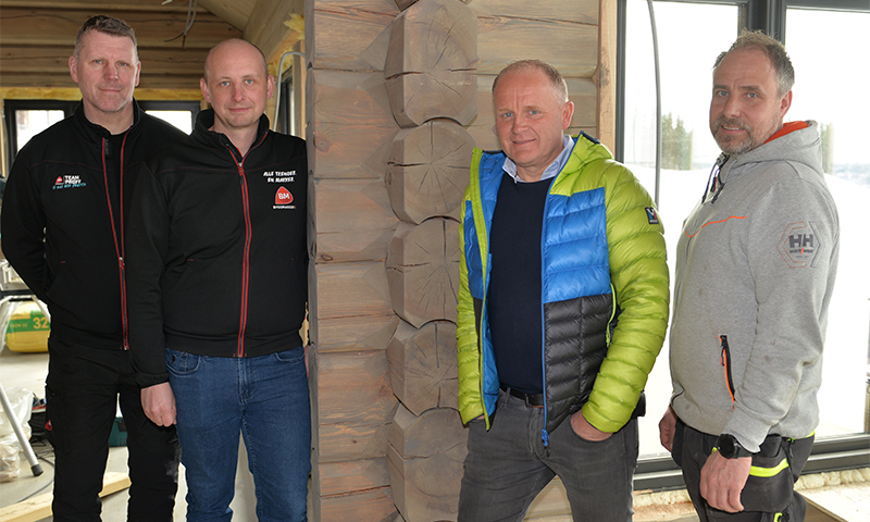 Fire menn i svarte, blå og grå jakker står foran tømmervegg i en snødekt hytte