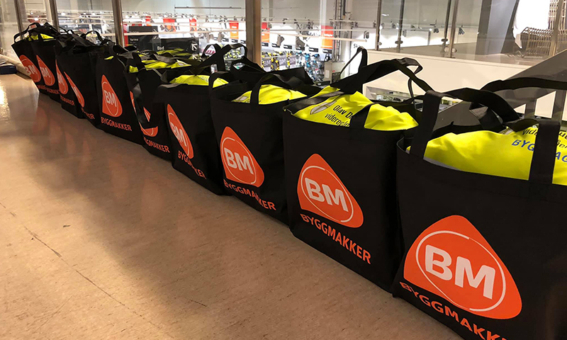 Rekke svarte Byggmakker-vesker med oransje BM-logo og gule refleksvester på butikkhylle