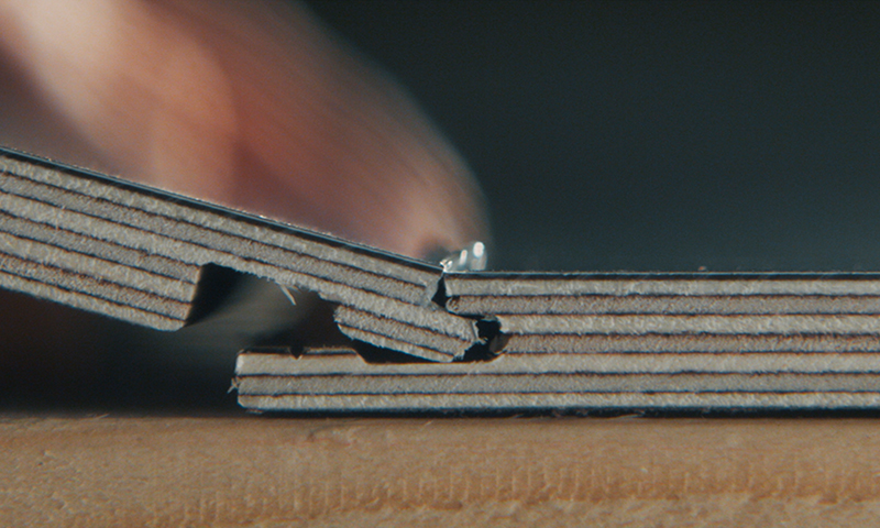 Nærbilde av lagdelt brun papp som bøyes ved hengsel, finger mot mørk bakgrunn