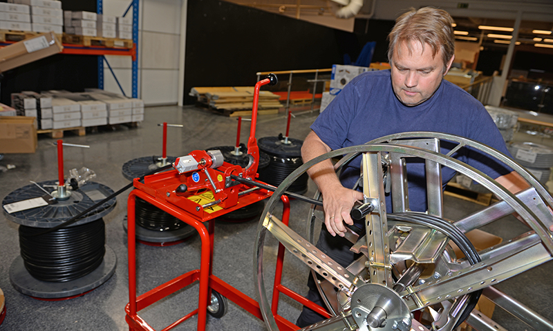 Person i blå skjorte mater svart kabel på rød kabeltrekker ved kabeltromler i lagerhall