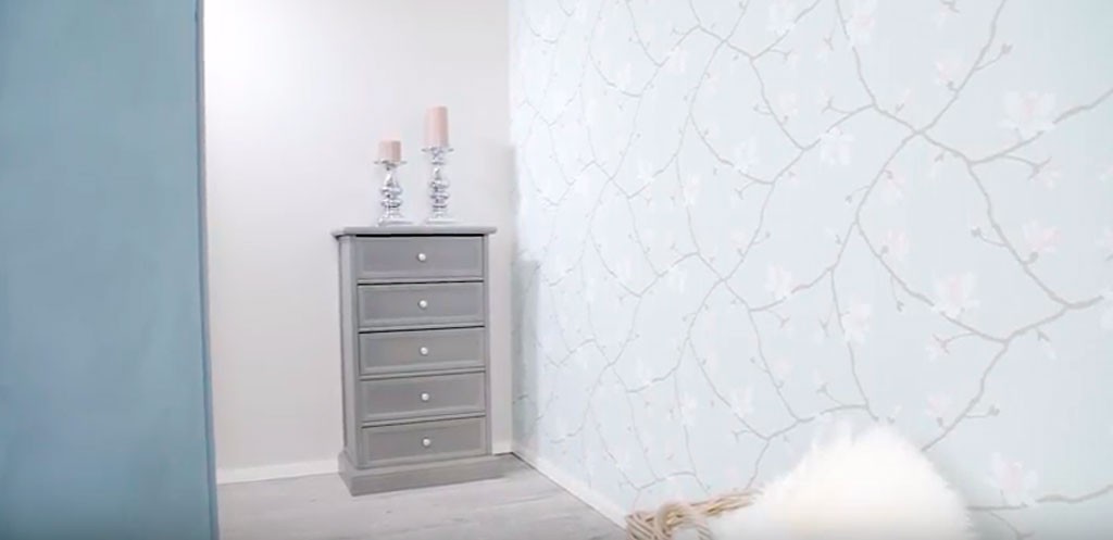Lysegrå kommode med fem skuffer og rosa lys på toppen, foran blomstrete tapet og hvit pelskurv.