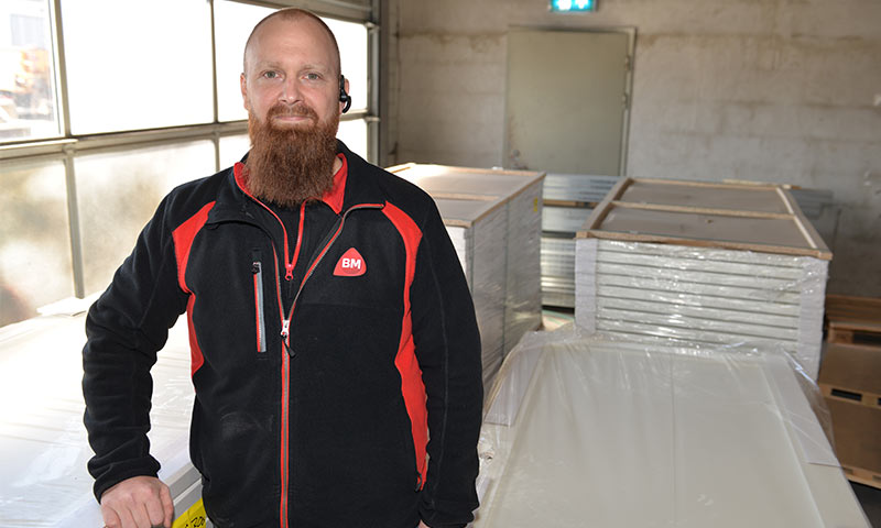 Ansatt i svart-rød BM-jakke står foran stabler med plastinnpakkede hvite paneler i lagerhall