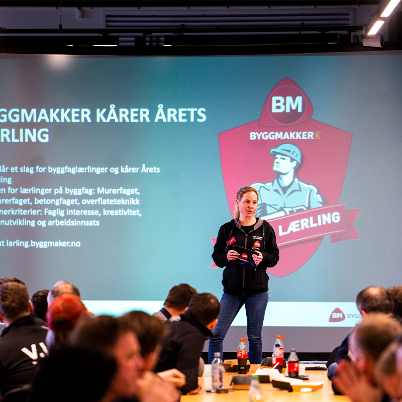 Person på scene foran gråblå skjerm med rødt Byggmakker-logo og Årets lærling-tekst.