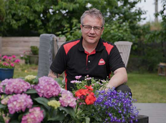 Mann i svart-rød skjorte ved bord med rosa hortensia, røde pelargonia og blå lobelia i hage.
