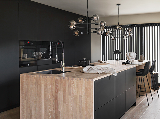 Moderne svart kjøkken med lys tremarbordsøy, glasspendler, barstoler og sorte vertikalpersienner