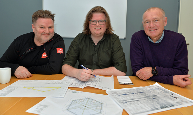 Tre personer ved lyst bord foran blågrå vegg med tekniske tegninger, en skriver med penn.