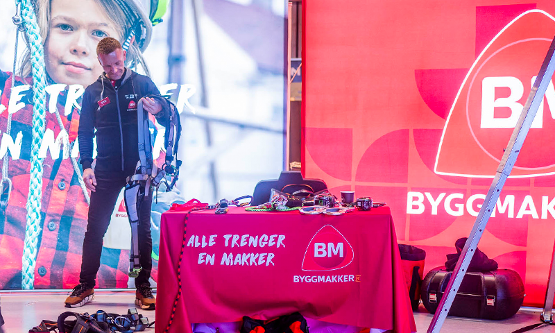 Stand med rødt Byggmakker-bord dekket med sikkerhetsutstyr og person holder fallsikringssele