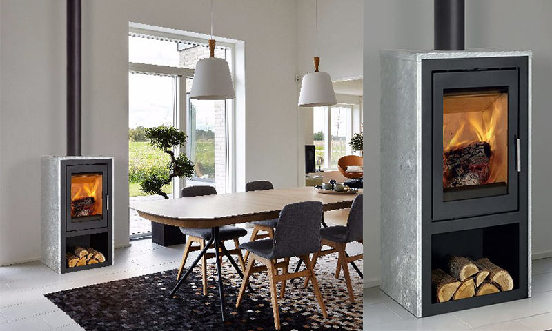 Hvit spisestue med trebord, grå stoler og steinovn med glødende flammer ved store vinduer.