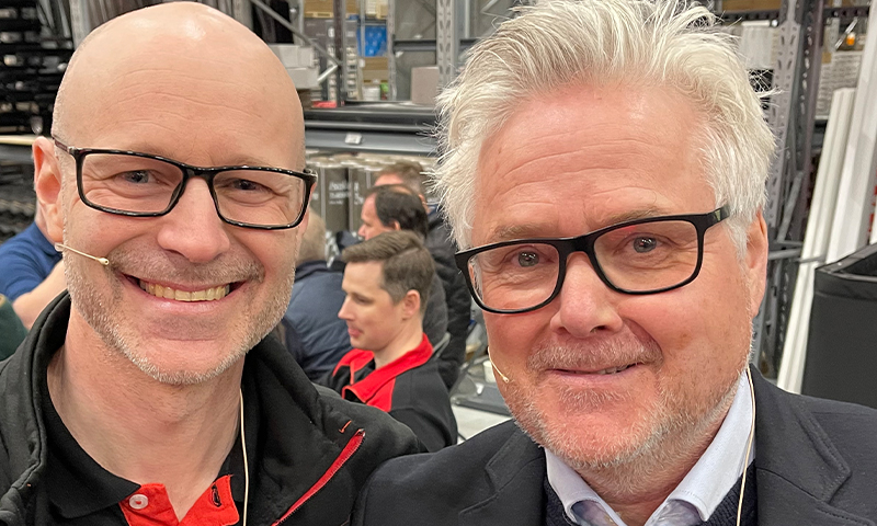 To menn i svarte og røde jakker står i travelt varehus med høye metallreoler.