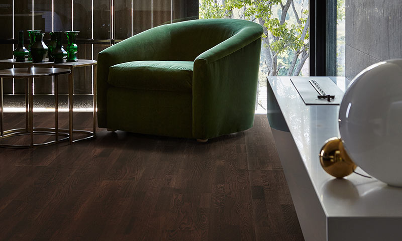 Grønn fløyelslenestol ved vindu med utsikt til trær, mørkt tregulv og små bord.